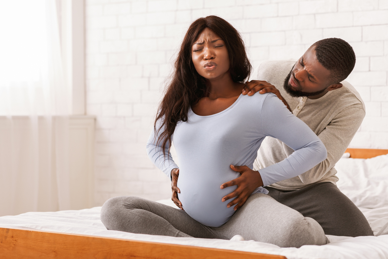 Pregnant black lady suffering from back pain, sitting on bed, her attentive husband massaging her shoulders, free space, Partners Can Help Their Person Go Through Childbirth