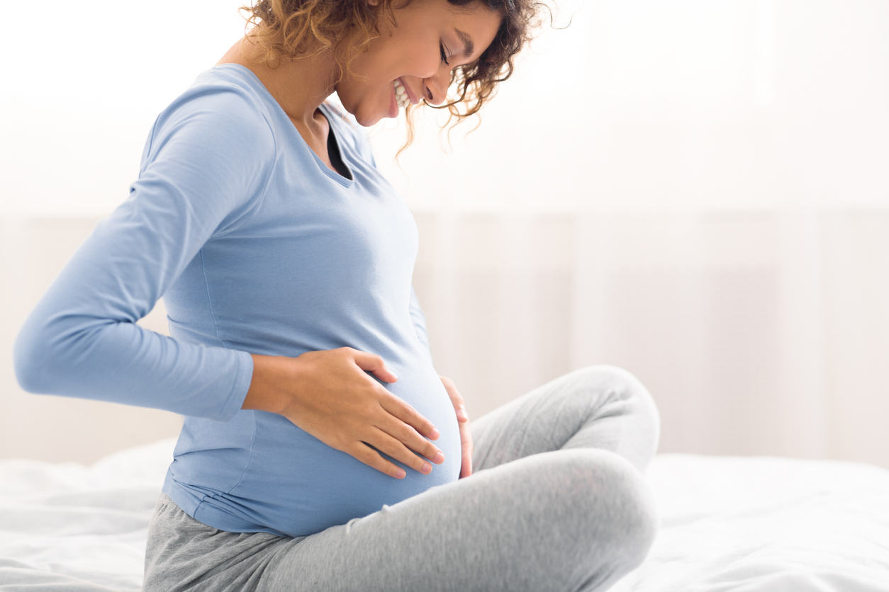Happy expectant woman touching belly, enjoying unborn baby pushings in bed, side view, wondering how pregnancy can impact your skin.