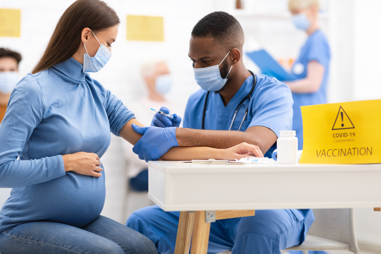 Covid-19 Vaccination Concept. Pregnant Woman Receiving Coronavirus Vaccine Intramuscular Injection Shot In Arm, Sitting With Black Nurse In Hospital Room. Covid 19 Virus Protection Medical Campaign.