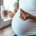 Pregnant woman at home taking a daily prenatal vitamin with a glass of water.