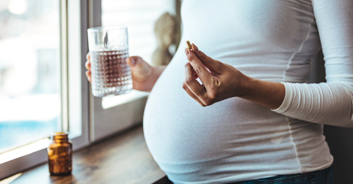 Pregnant woman at home taking a daily prenatal vitamin with a glass of water.